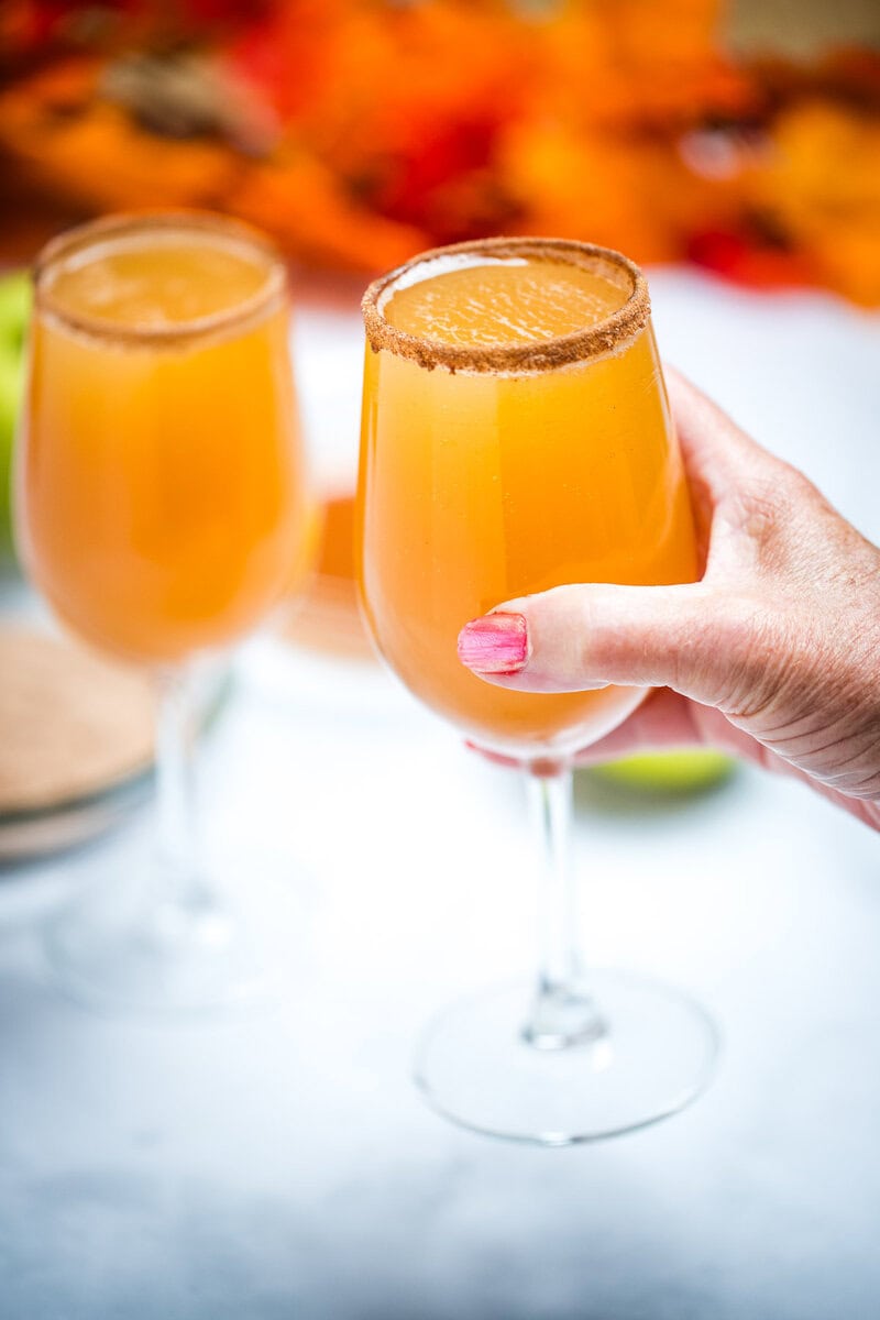 A hand holding a wine glass filled with apple cider mimosas with a cinnamon sugar rim, with another filled wine glass on a table in the background. Behind the glasses there is a shallow bowl with cinnamon sugar, green apples, and fall leaves.