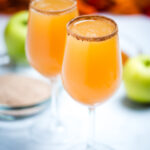 A wine glass filled with apple cider mimosas with a cinnamon sugar rim, with another filled wine glass on a table in the background. Behind the glasses there is a shallow bowl with cinnamon sugar, green apples, and fall leaves.