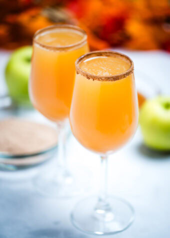 A wine glass filled with apple cider mimosas with a cinnamon sugar rim, with another filled wine glass on a table in the background. Behind the glasses there is a shallow bowl with cinnamon sugar, green apples, and fall leaves.