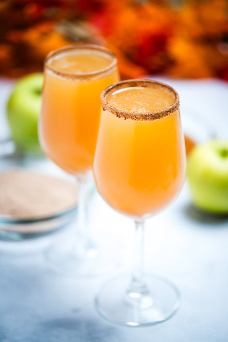 A wine glass filled with apple cider mimosas with a cinnamon sugar rim, with another filled wine glass on a table in the background. Behind the glasses there is a shallow bowl with cinnamon sugar, green apples, and fall leaves.