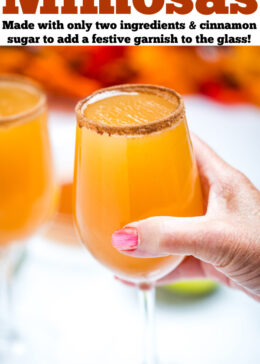 Pinterest pin with a hand holding a wine glass filled with apple cider mimosas, with another wine glass on a table in the background.