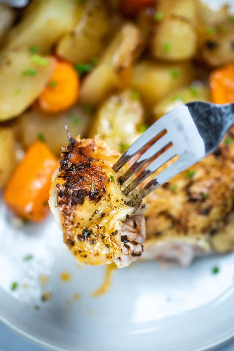 A fork lifting a piece of roasted chicken with caramelized skin and herbs above a plate with a chicken breast, potatoes, and carrots topped with chopped chives.