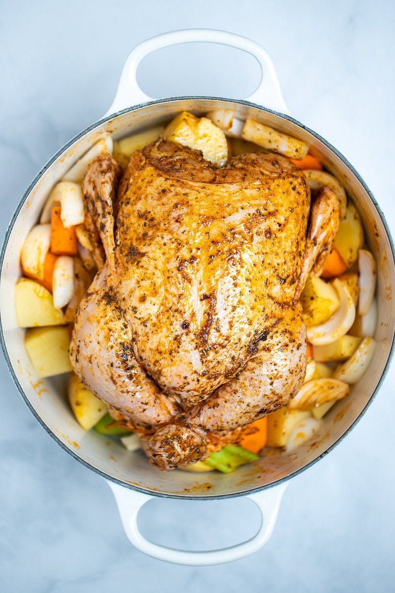 A dutch oven full of uncooked carrots, celery, and potatoes with a raw chicken on top, rubbed with olive oil, herbs, and spices before being cooked.