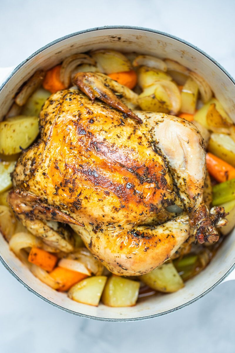 A dutch oven full of cooked carrots, celery, and potatoes with a roasted chicken on top, rubbed with olive oil, herbs, and spices before being served.