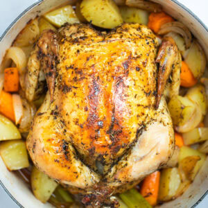 A dutch oven full of cooked carrots, celery, and potatoes with a roasted chicken on top, rubbed with olive oil, herbs, and spices before being served.