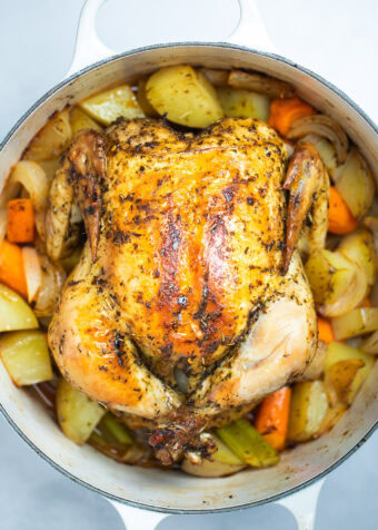 A dutch oven full of cooked carrots, celery, and potatoes with a roasted chicken on top, rubbed with olive oil, herbs, and spices before being served.