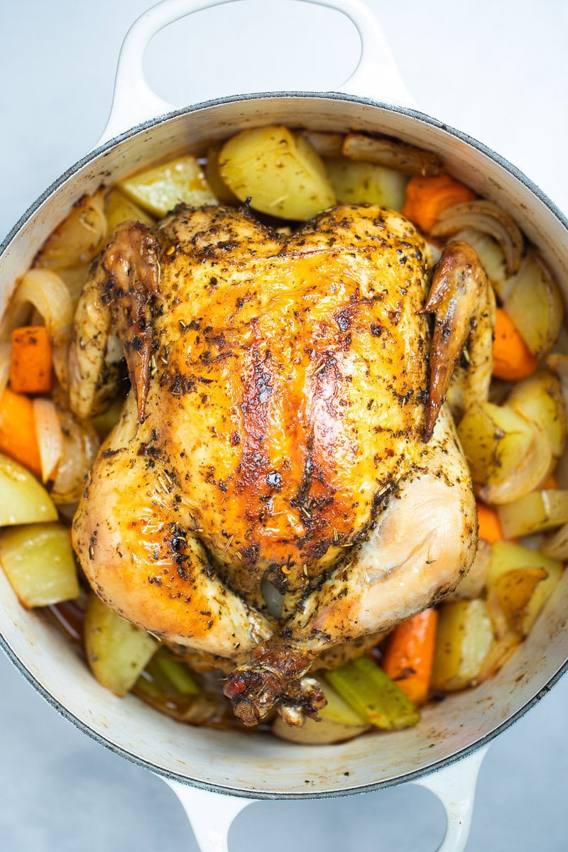 A dutch oven full of cooked carrots, celery, and potatoes with a roasted chicken on top, rubbed with olive oil, herbs, and spices before being served.