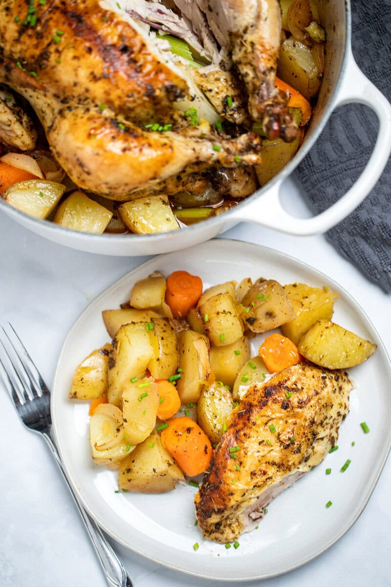 A roasted chicken breast and potatoes topped with chopped chives on a plate next to a fork, in front of a dutch oven with a whole chicken on a table.