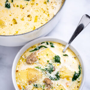 A bowl of sausage potato soup with a spoon in it topped with parmesan cheese, in front of a dutch oven of soup, on a table.