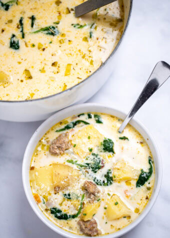 A bowl of sausage potato soup with a spoon in it topped with parmesan cheese, in front of a dutch oven of soup, on a table.