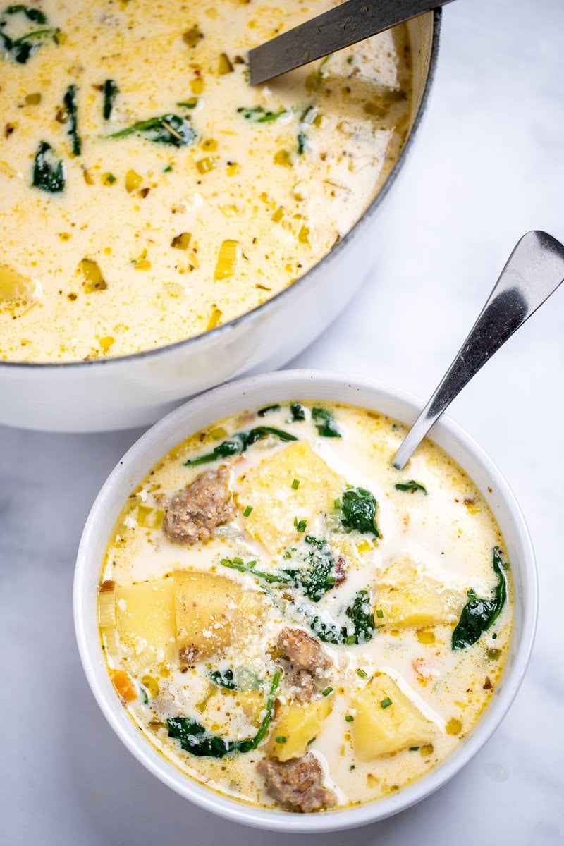 A bowl of sausage potato soup with a spoon in it topped with parmesan cheese, in front of a dutch oven of soup, on a table.