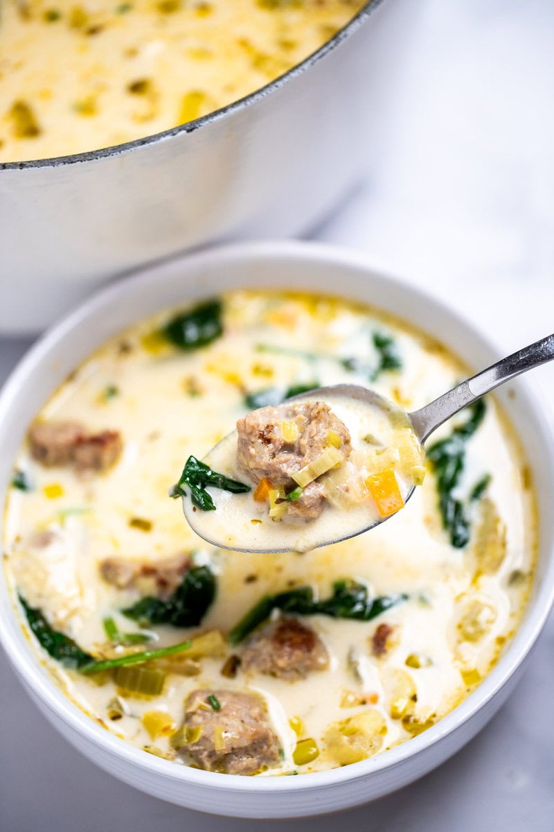 A bowl of sausage potato soup in front of a dutch oven of soup, with a spoon lifting some soup up with a big piece of sausage on top.