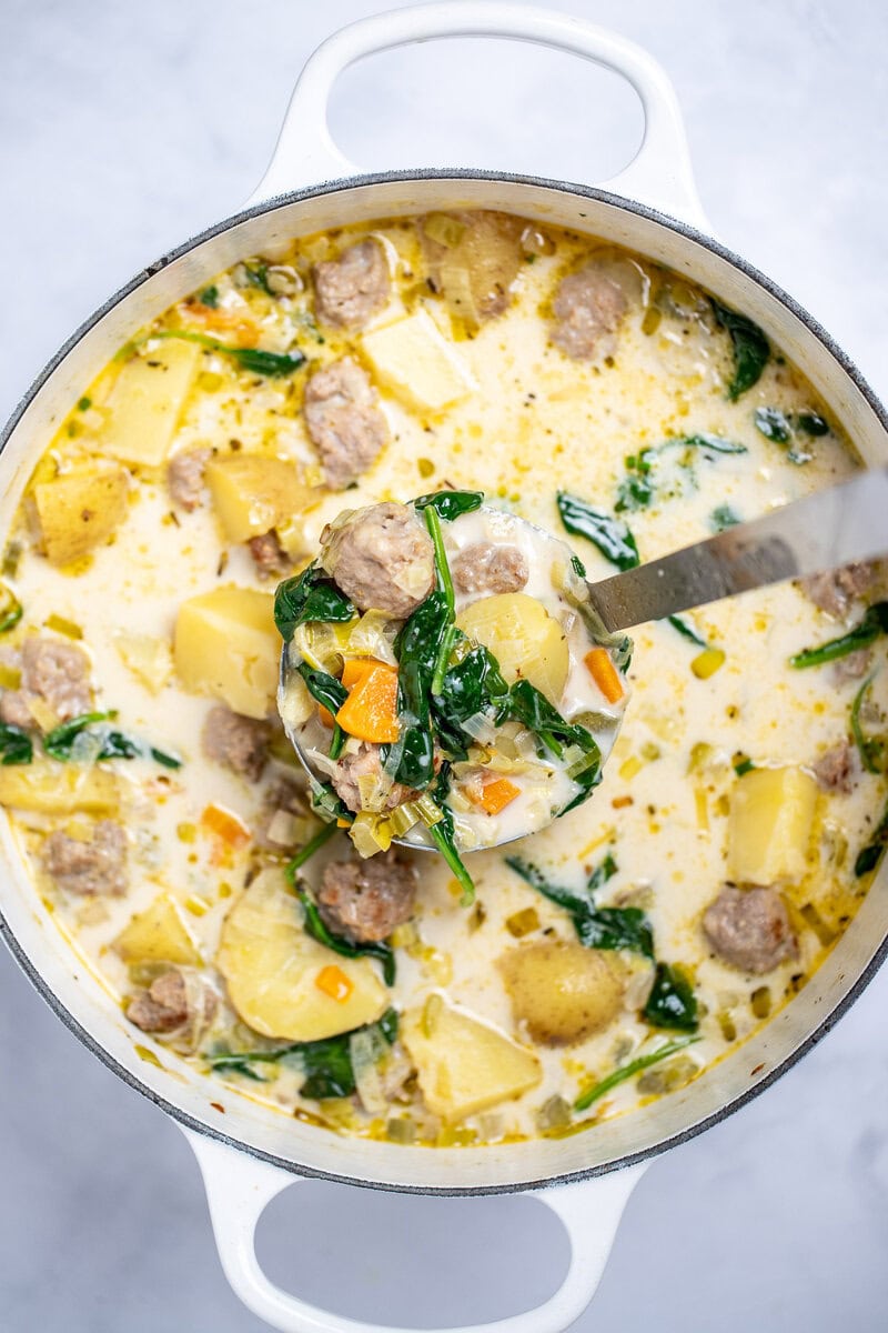 A dutch oven of sausage potato soup on a table, with a ladle lifting some soup out of the pot.