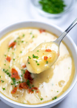 A bowl of slow cooker potato soup topped with chives and crispy bacon, with a spoon lifting up some soup, and a bowl of chives on the table in the background.