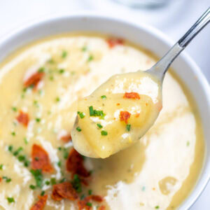 A bowl of slow cooker potato soup topped with chives and crispy bacon, with a spoon lifting up some soup, and a bowl of chives on the table in the background.