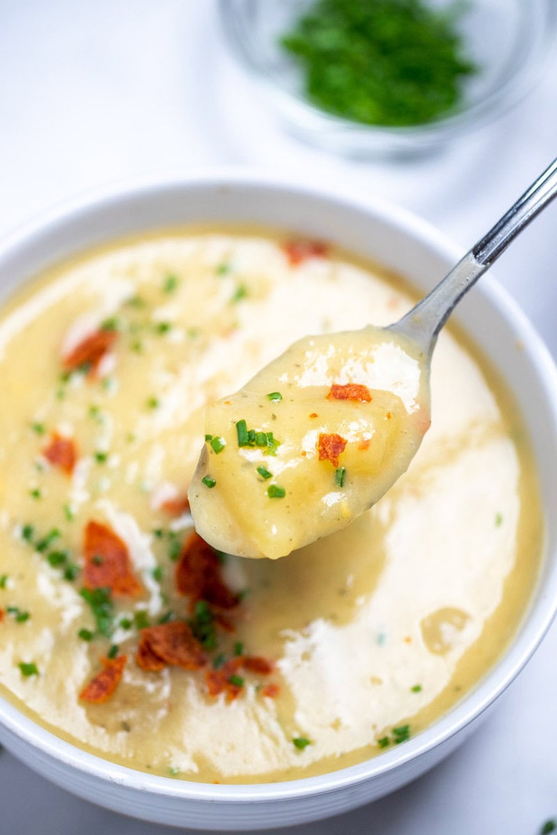 A bowl of slow cooker potato soup topped with chives and crispy bacon, with a spoon lifting up some soup, and a bowl of chives on the table in the background.