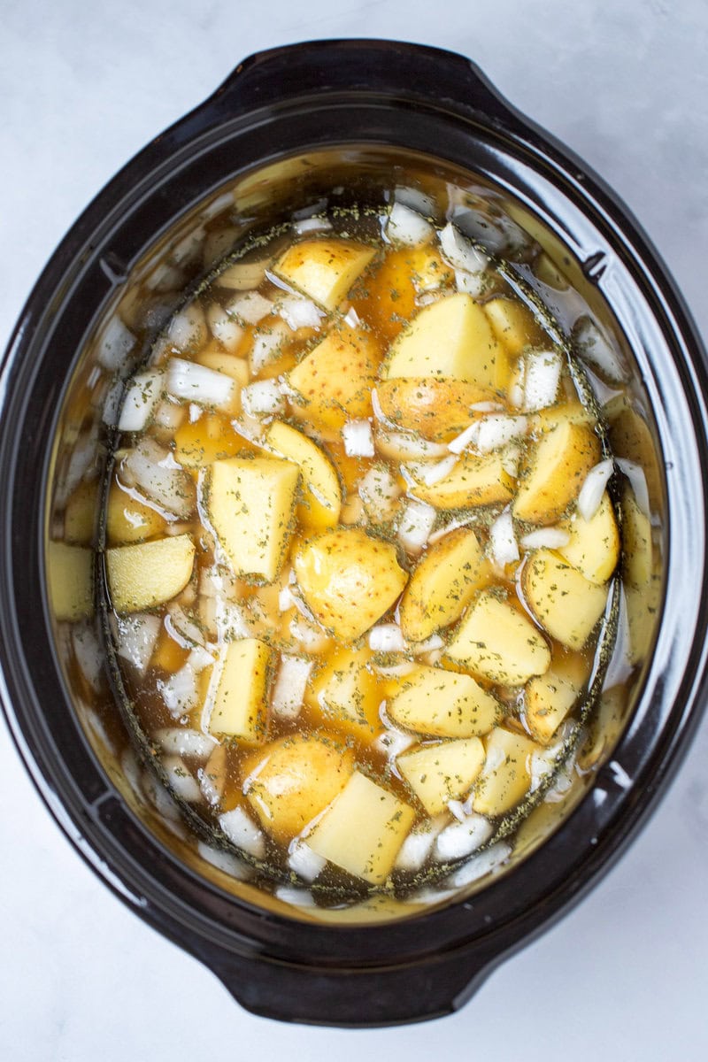 A slow cooker with potato pieces, onion, seasoning, and broth before being cooked.