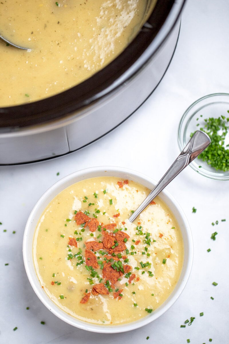 A bowl of slow cooker potato soup topped with chives and crispy bacon, with a spoon in the soup, and a bowl of chives and a slow cooker on the table in the background.