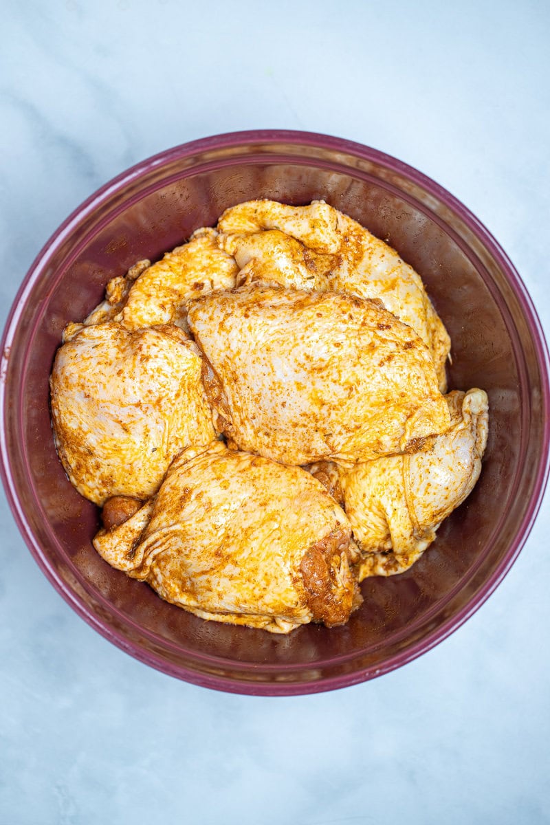 A mixing bowl on a table full of raw chicken thighs being coated with olive oil and spices.