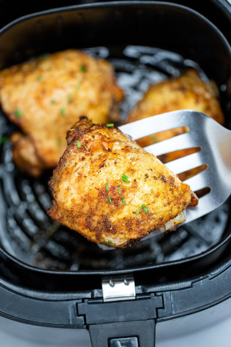 Chicken thighs in an air fryer basket with chopped chives on top of crispy skin, with a spatula lifting one up.