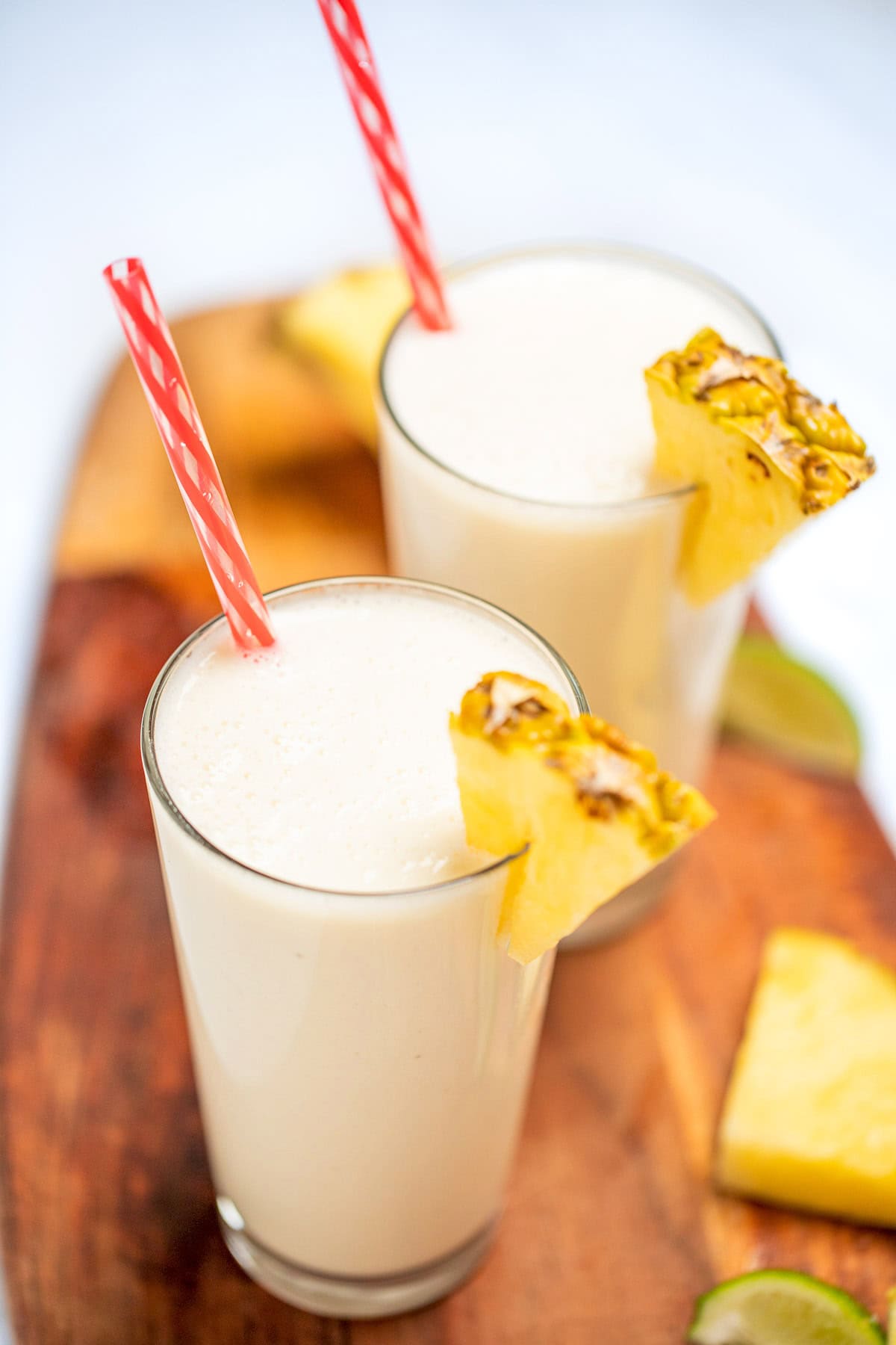 A tall glass with a pineapple wedge garnish and a straw, full of pina colada smoothie, with another glass behind it, on a cutting board with pineapple and lime wedges.