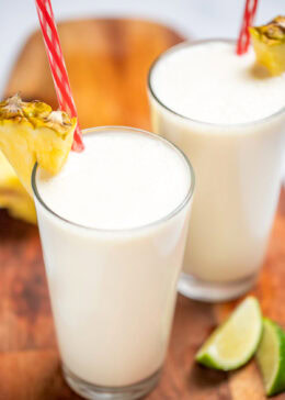 A tall glass with a pineapple wedge garnish and a straw, full of pina colada smoothie, with another glass behind it, on a cutting board with pineapple and lime wedges.