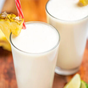 A tall glass with a pineapple wedge garnish and a straw, full of pina colada smoothie, with another glass behind it, on a cutting board with pineapple and lime wedges.