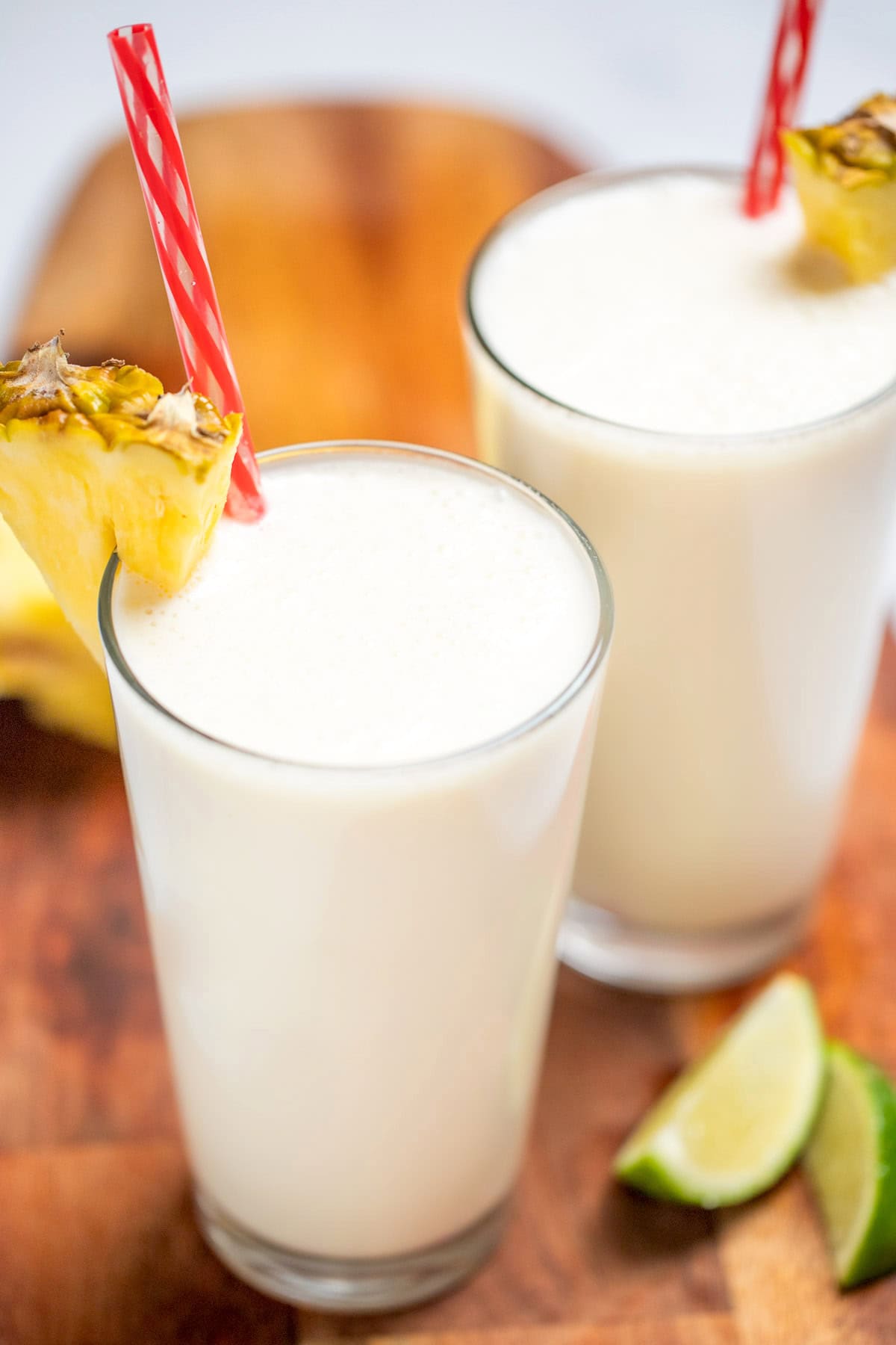 A tall glass with a pineapple wedge garnish and a straw, full of pina colada smoothie, with another glass behind it, on a cutting board with pineapple and lime wedges.