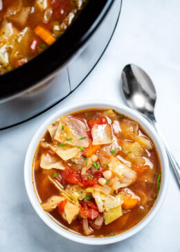 A bowl of slow cooker cabbage soup on the table, with a spoon next to the soup, in front of a slow cooker.