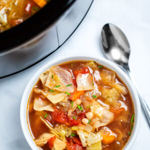 A bowl of slow cooker cabbage soup on the table, with a spoon next to the soup, in front of a slow cooker.