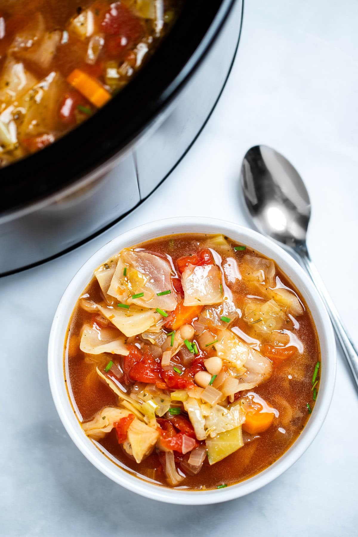 A bowl of slow cooker cabbage soup on the table, with a spoon next to the soup, in front of a slow cooker.