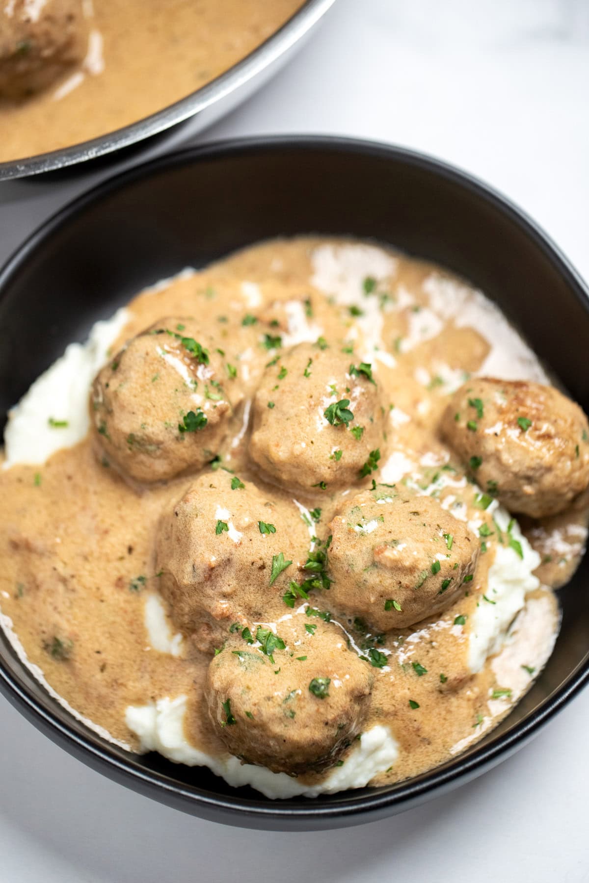 A bowl with meatballs in gravy topped with fresh parsley, on top of mashed potatoes, in front of a skillet.