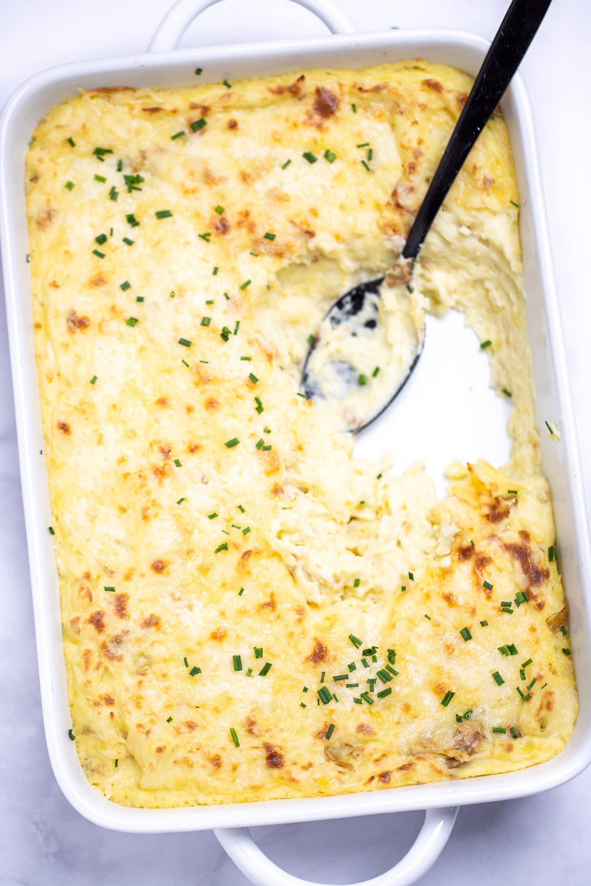 A casserole dish full of twice baked mashed potatoes with caramelized cheese on top with fresh chopped chives, with a spoon resting in an empty spot in the dish.