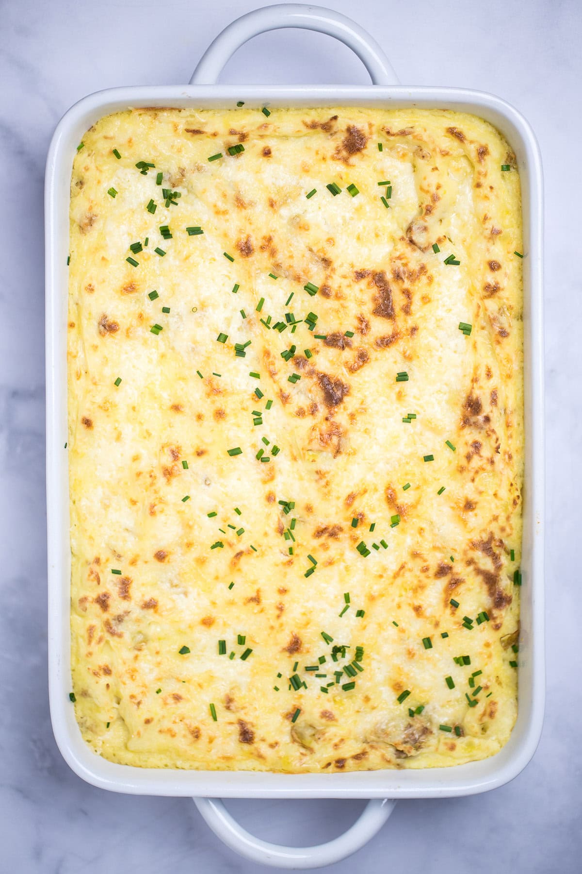 A casserole dish full of twice baked mashed potatoes with caramelized cheese on top with fresh chopped chives, on a table.