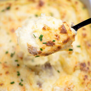 A casserole dish full of twice baked mashed potatoes with caramelized cheese on top with fresh chopped chives, with a spoon lifting some mashed potatoes up.