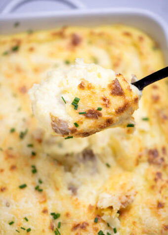 A casserole dish full of twice baked mashed potatoes with caramelized cheese on top with fresh chopped chives, with a spoon lifting some mashed potatoes up.