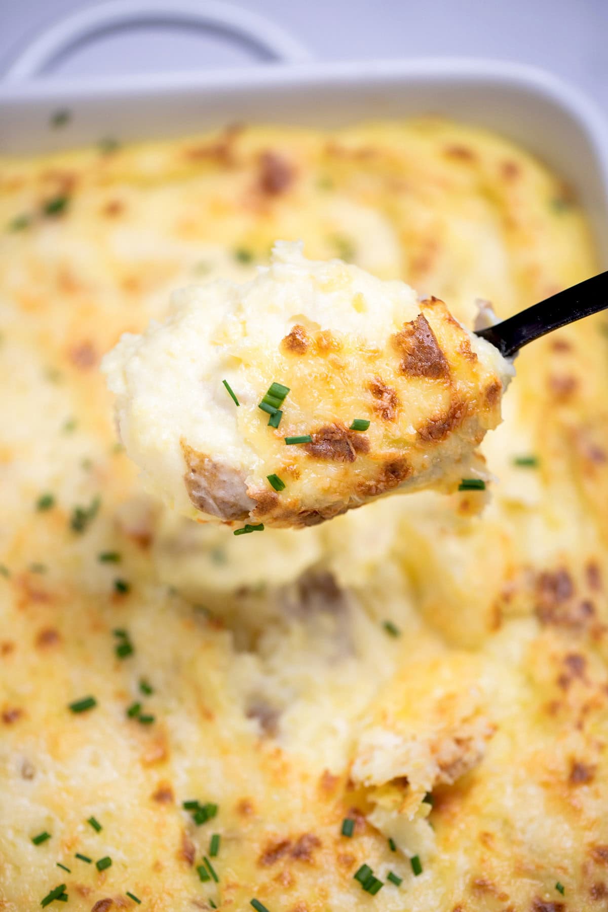 A casserole dish full of twice baked mashed potatoes with caramelized cheese on top with fresh chopped chives, with a spoon lifting some mashed potatoes up.