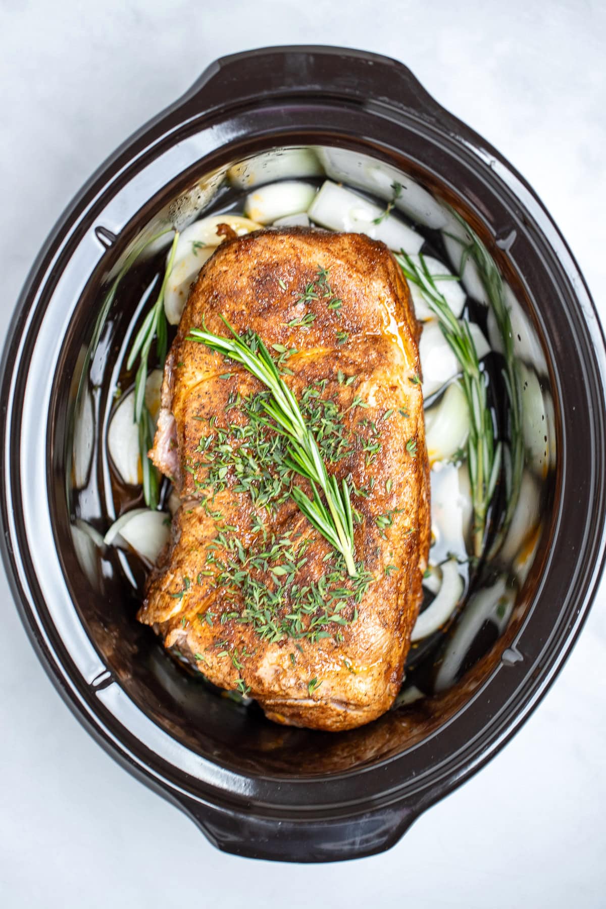 An uncooked pork roast that's been covered in seasoning and seared, in a slow cooker sitting on top of raw onions and broth, and topped with fresh thyme and rosemary.