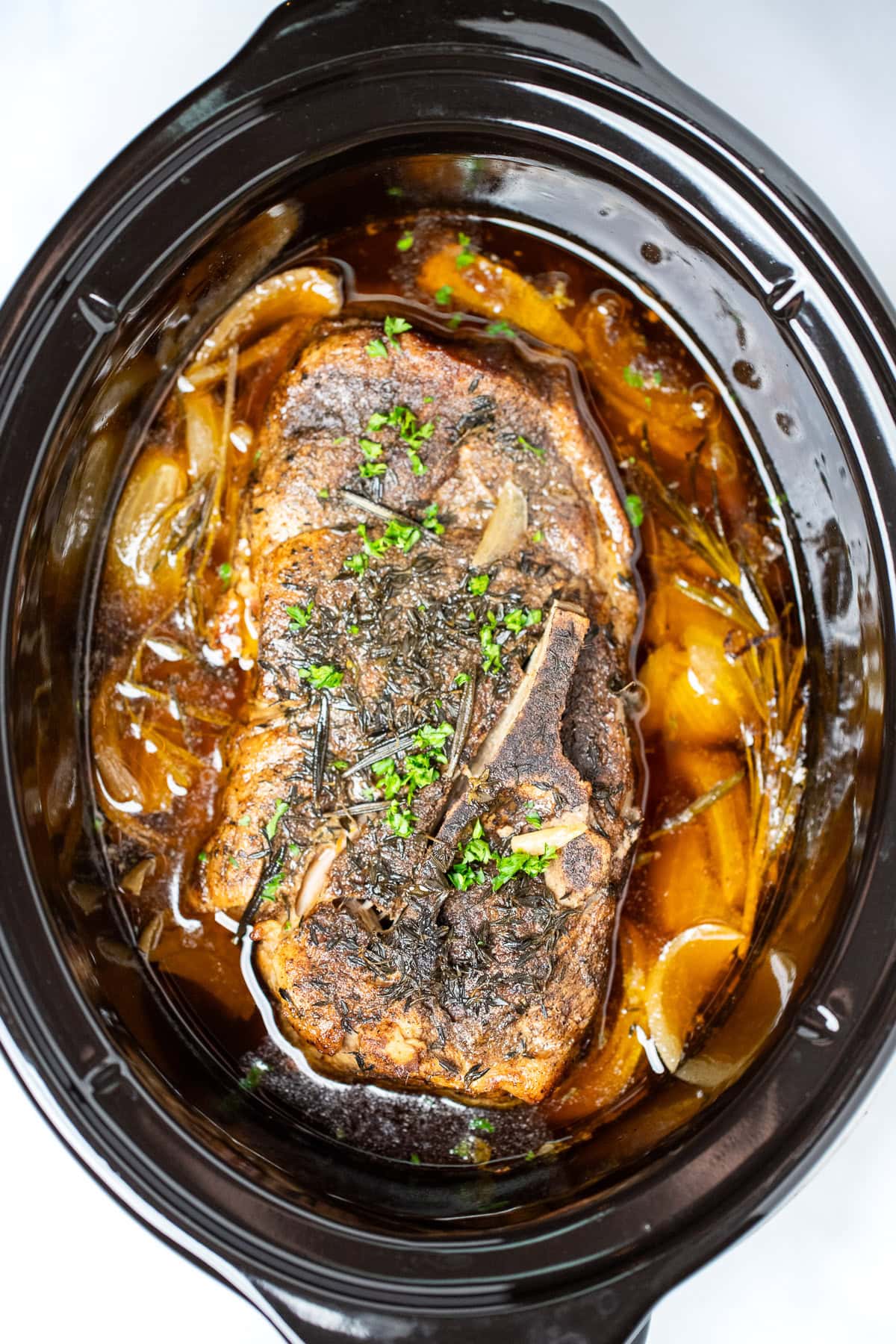 A fully cooked pork roast in a slow cooker with broth and onions, topped with fresh parsley.