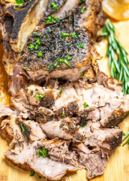 A pork roast on a cutting board topped with fresh parsley, next to fresh rosemary, with the front part of the roast shredded.