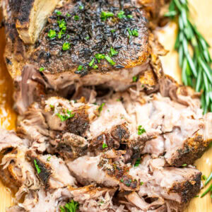 A pork roast on a cutting board topped with fresh parsley, next to fresh rosemary, with the front part of the roast shredded.