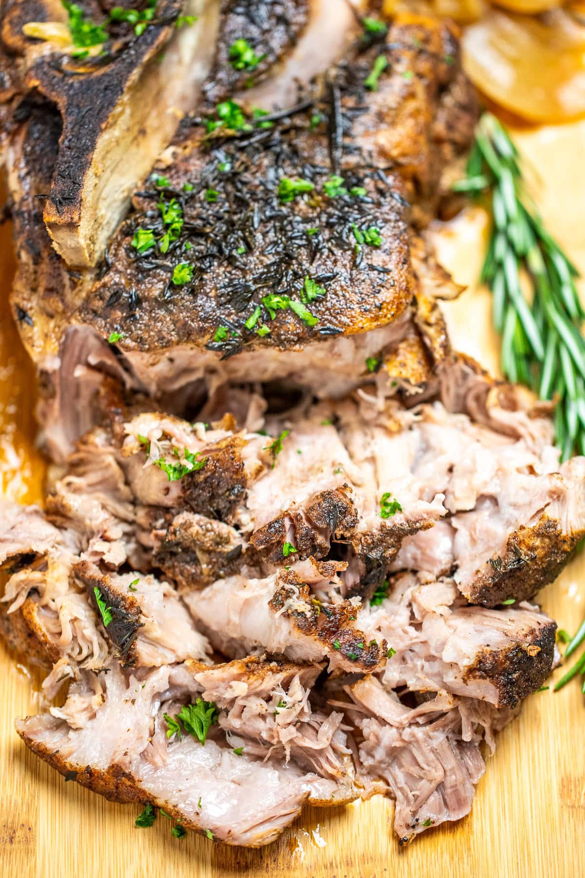 A pork roast on a cutting board topped with fresh parsley, next to fresh rosemary, with the front part of the roast shredded.