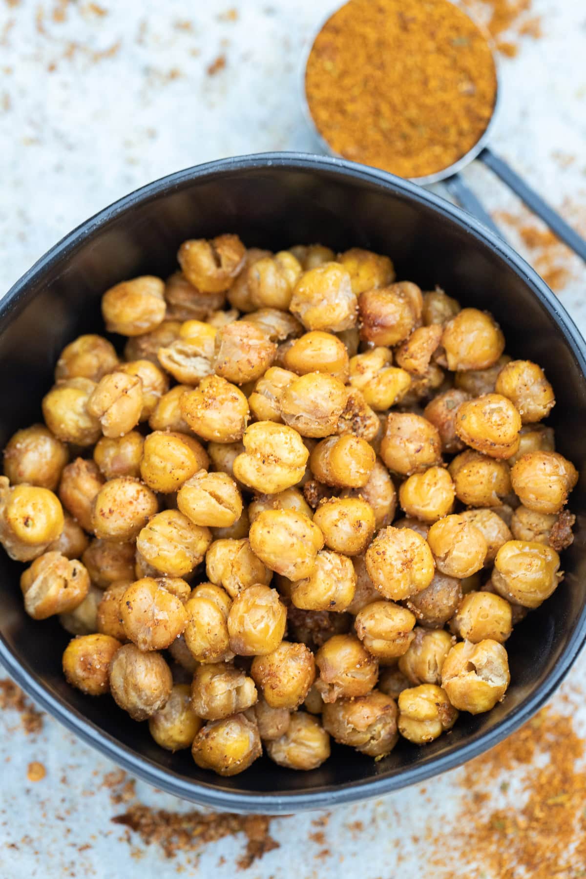 A bowl on a table with crispy air fryer chickpeas coated in seasoning, with a measuring cup of spices in the background.