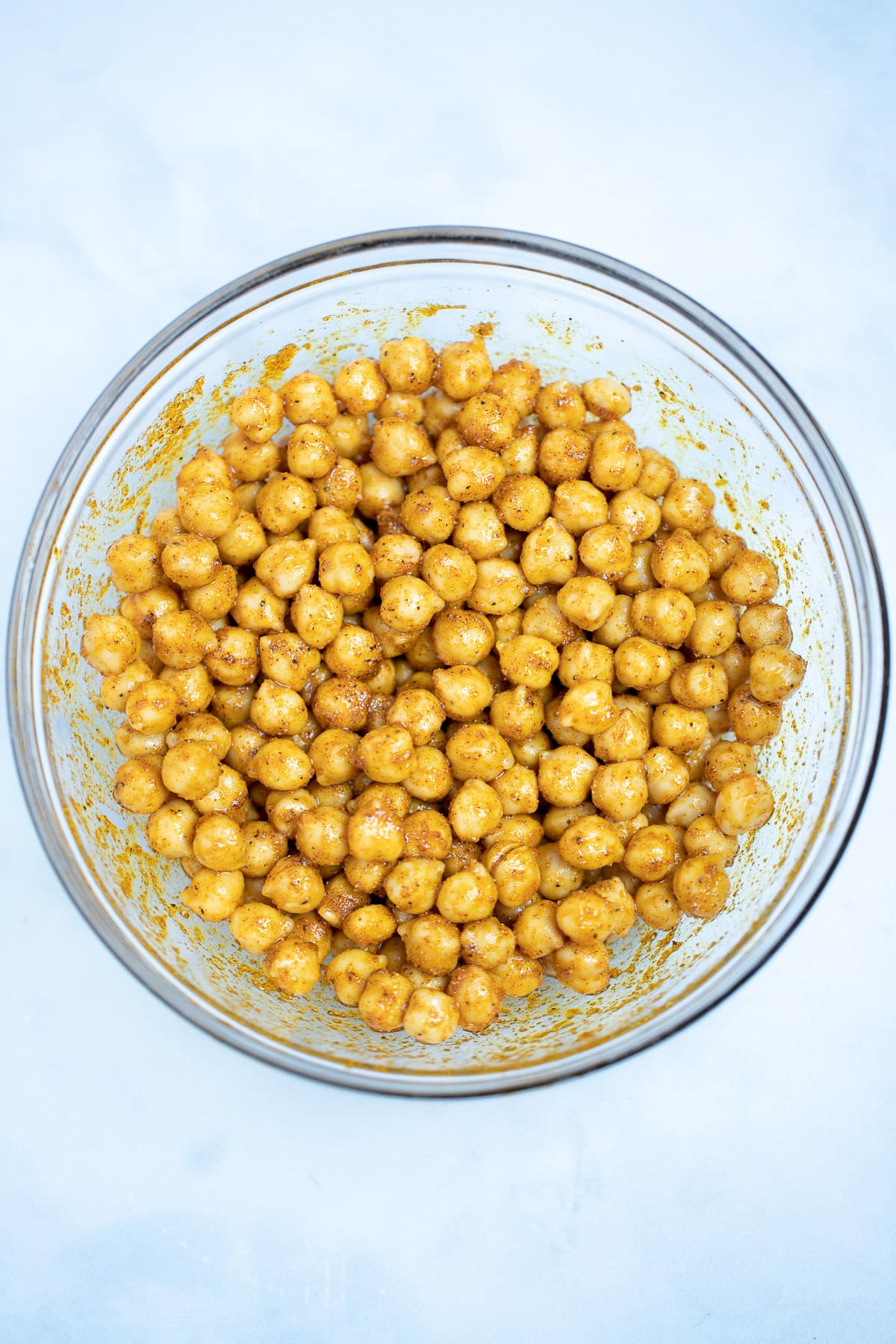 A glass mixing bowl with chickpeas coated in olive oil and seasoning on a table.