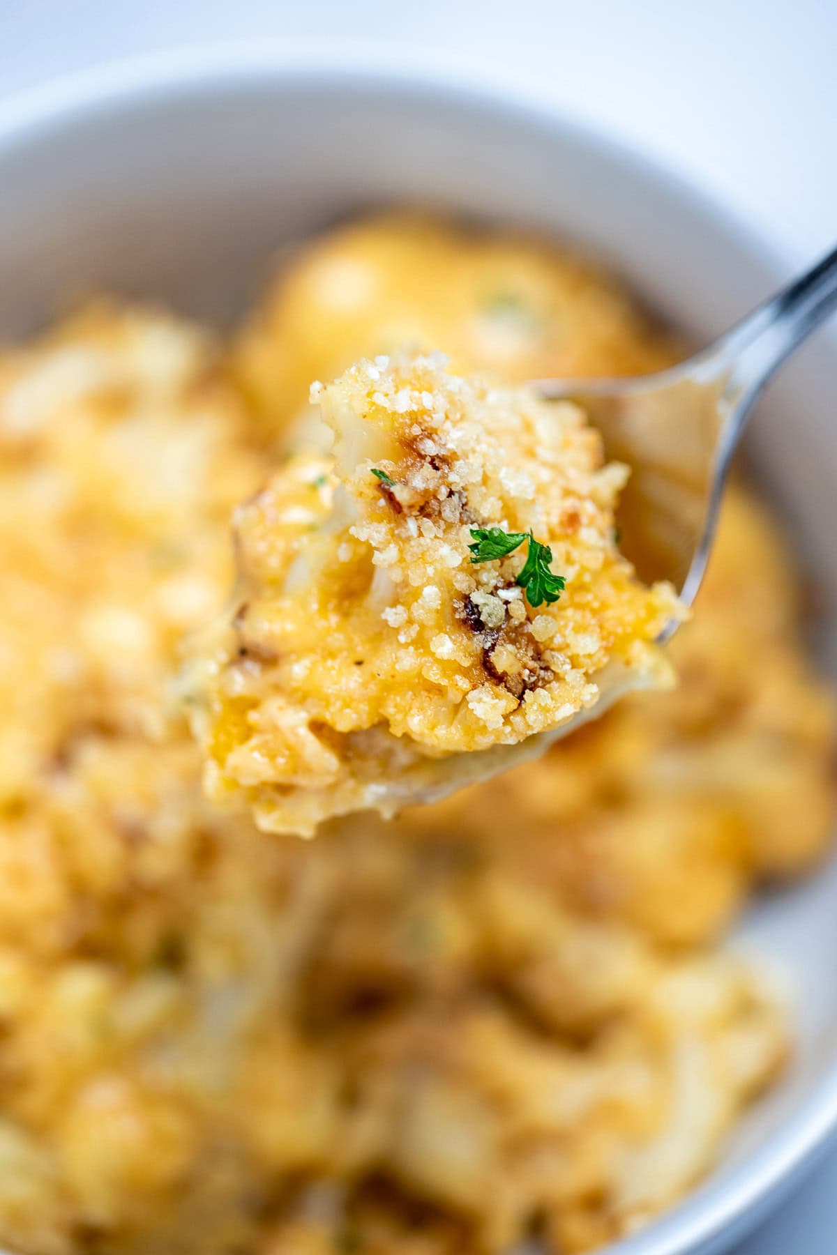 A spoon lifting up cauliflower mac and cheese from a bowl, topped with fresh chopped parsley.