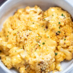 A bowl full of cauliflower mac and cheese on a table next to a spoon and a baking dish.