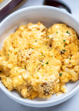 A bowl full of cauliflower mac and cheese on a table next to a spoon and a baking dish.