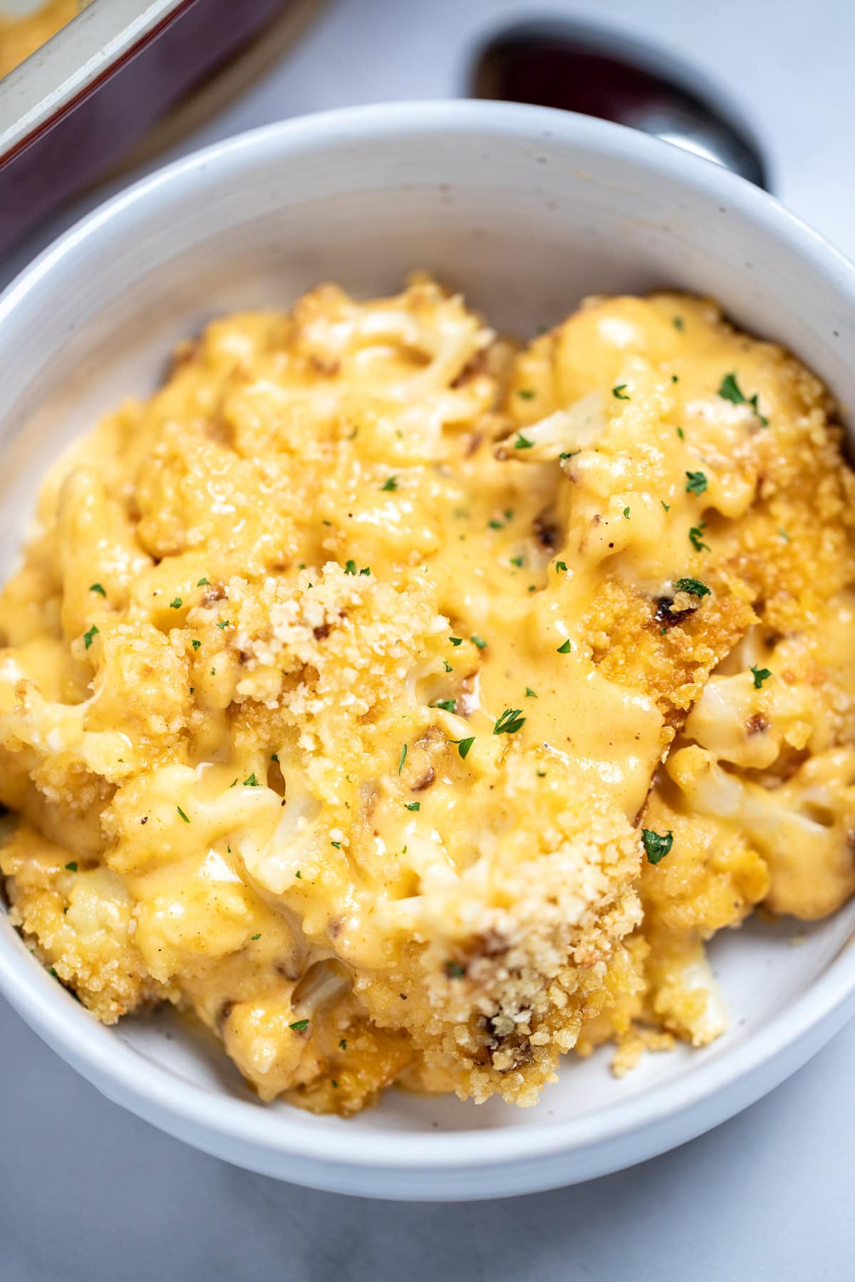 A bowl full of cauliflower mac and cheese on a table next to a spoon and a baking dish.