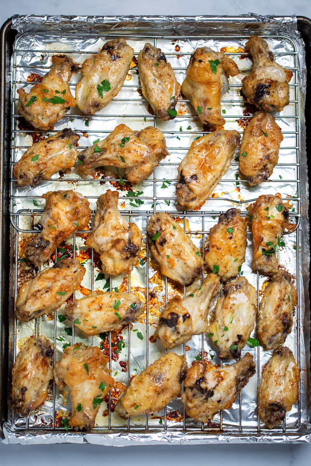 A sheet pan lined with tinfoil and a baking rack with cooked lemon pepper chicken wings on it topped with lemon butter sauce and fresh parsley.