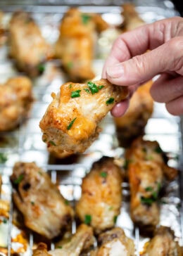 A hand holding a chicken wing above a baking rack of cooked chicken wings topped with fresh parsley.